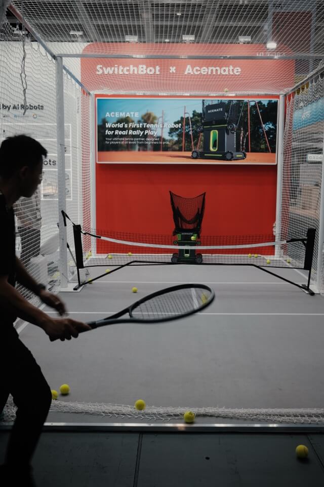 Ein Mann spielt in einem Netzgehege auf einer Messe Tennis gegen eine Ballmaschine.