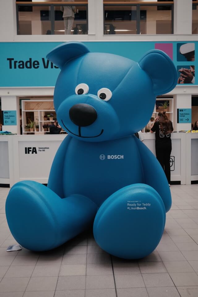 Ein großer, aufblasbarer, blauer Teddybär der Marke Bosch sitzt in einer Messehalle.
