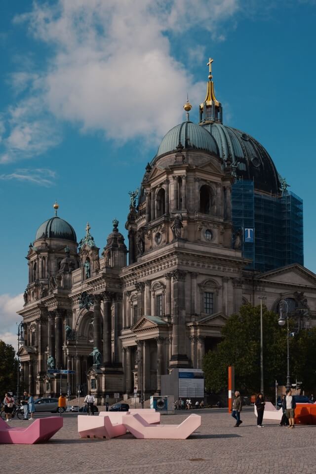Der Berliner Dom bei blauem Himmel mit modernen, rosa Sitzbänken auf dem Vorplatz.