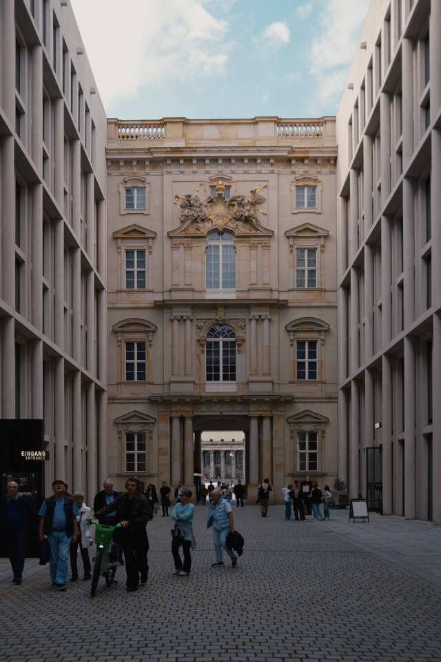 Menschen gehen durch einen gepflasterten Innenhof auf eine historische Fassade zu.