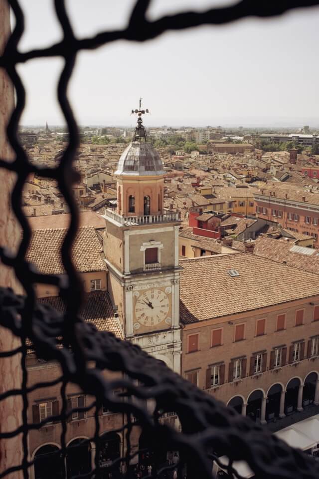 Der Blick durch ein dunkles Gitter auf einen Uhrturm über den Dächern von Modena.