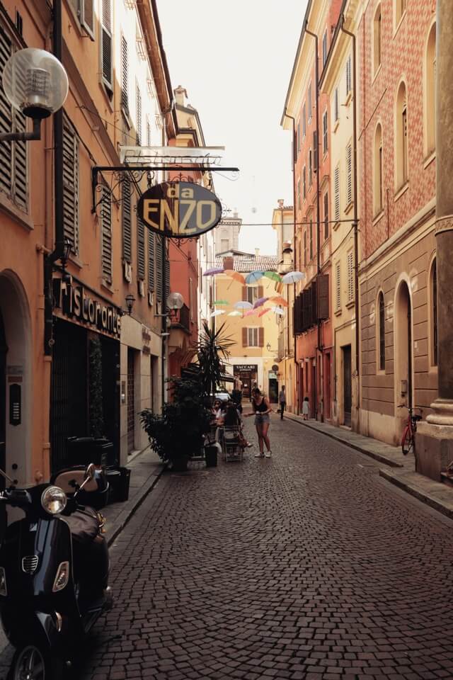 Eine belebte, enge Gasse in Italien mit Kopfsteinpflaster und bunten Regenschirmen in der Luft.