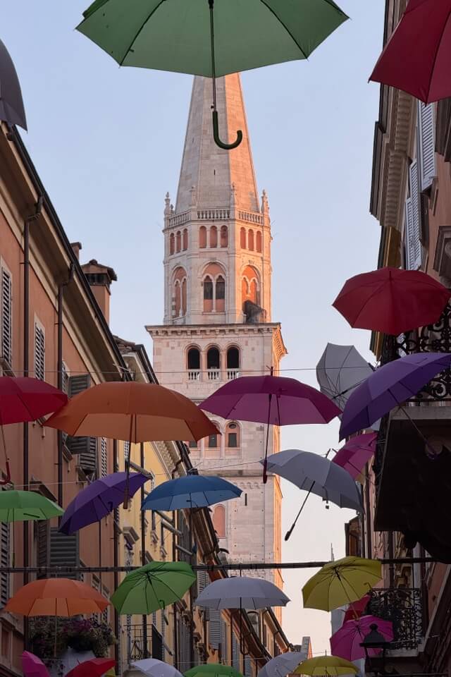 Viele bunte Regenschirme schweben in einer Gasse vor dem von der Sonne angestrahlten Glockenturm.
