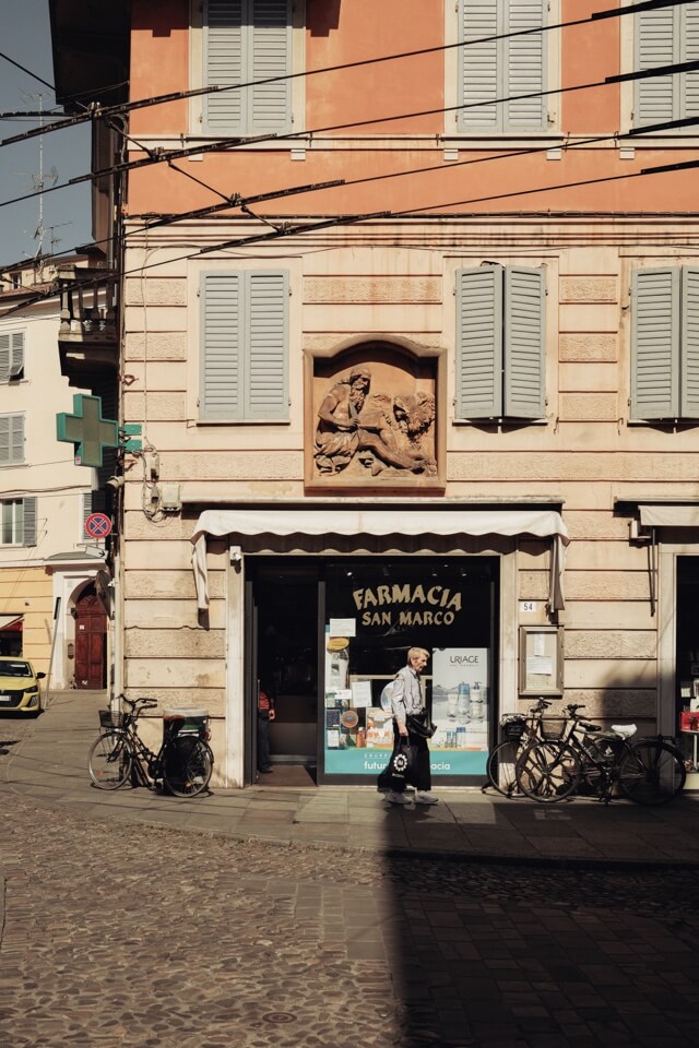 Eine ältere Person geht auf dem Kopfsteinpflaster an einer italienischen Apotheke vorbei.