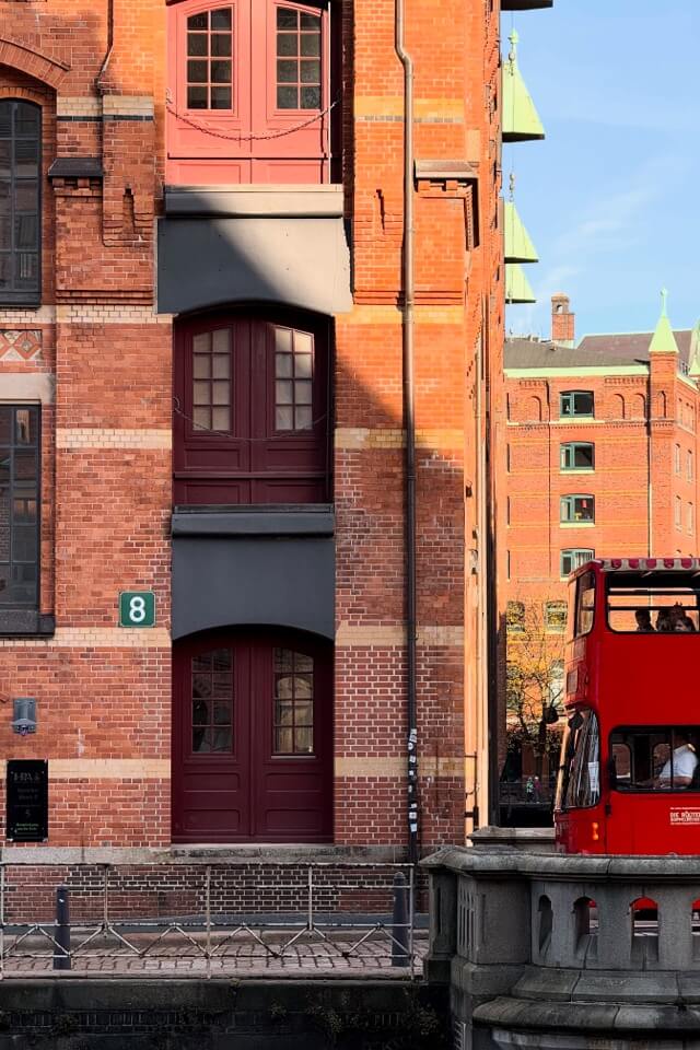 Ein roter Sightseeing-Bus fährt an einem sonnenbeschienenen Backstein-Speichergebäude in Hamburg vorbei.