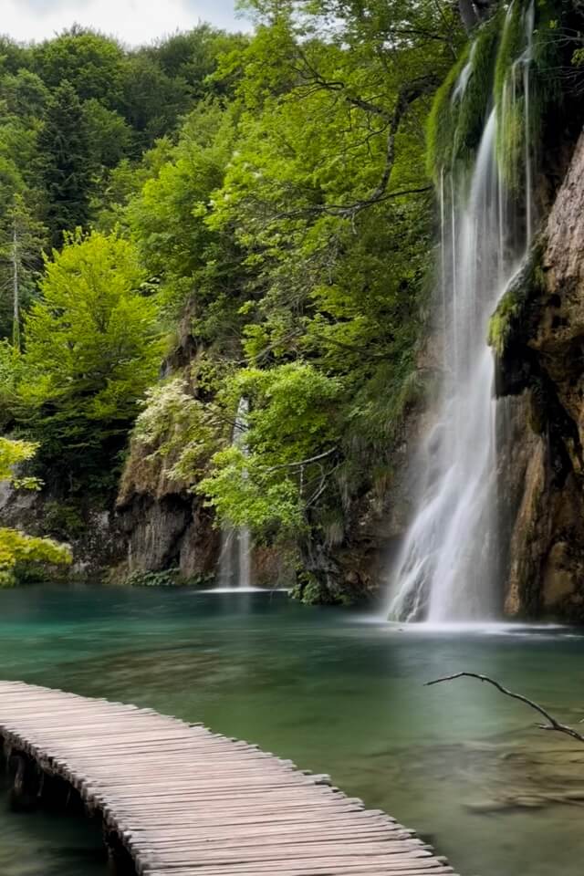 Ein Holzsteg verläuft am Ufer eines Sees entlang auf einen Wasserfall zu.