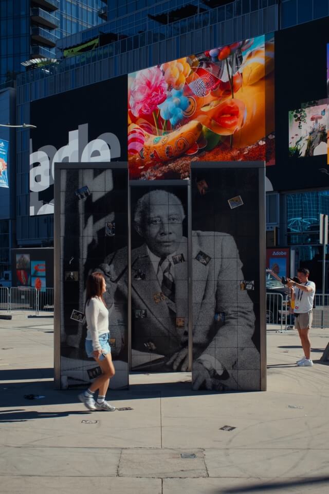 Passanten betrachten eine große Kunstinstallation mit einem Schwarz-Weiß-Porträt und bunten Grafiken auf einem städtischen Platz.