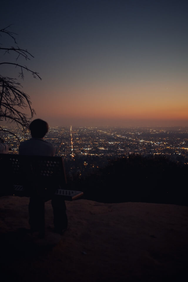 Eine Person sitzt auf einer Bank und blickt bei Dämmerung über das Lichtermeer von Los Angeles.