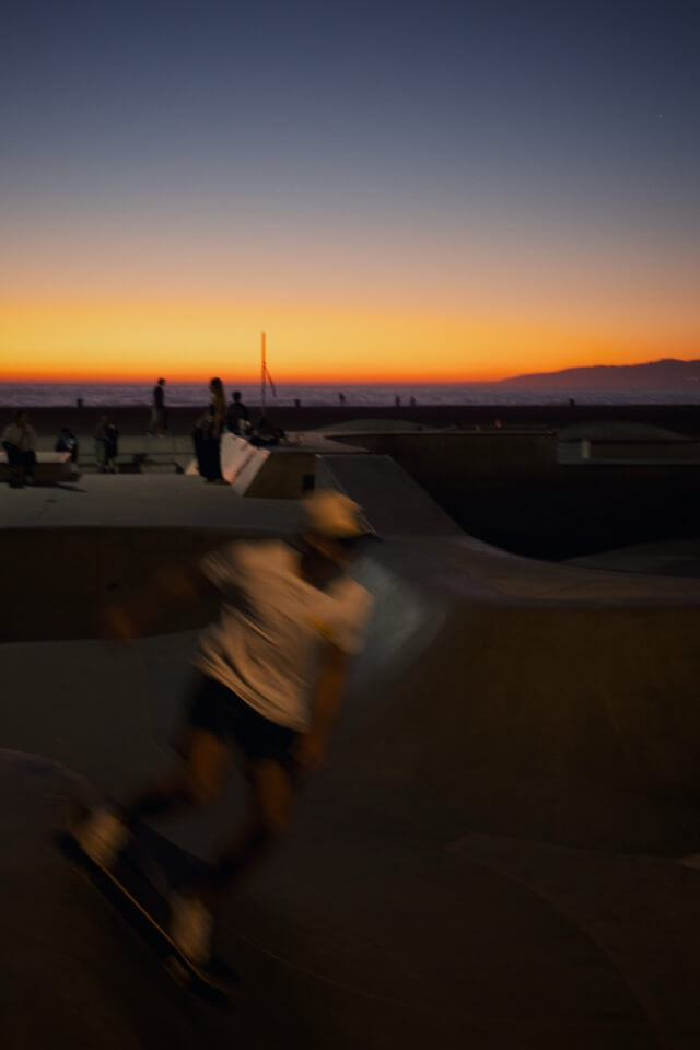 Ein Skateboarder in Bewegungsunschärfe fährt in einem Skatepark am Strand während eines leuchtenden orangefarbenen Sonnenuntergangs.