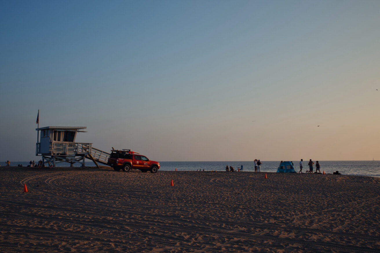 Ein Rettungsschwimmerturm und ein roter Pick-up-Truck der Rettungsschwimmer stehen bei Sonnenuntergang an einem weiten Sandstrand.