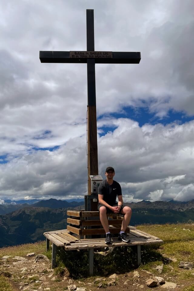 Ein junger Mann sitzt auf einer Holzbank unter einem großen Gipfelkreuz vor einem weiten Bergpanorama.
