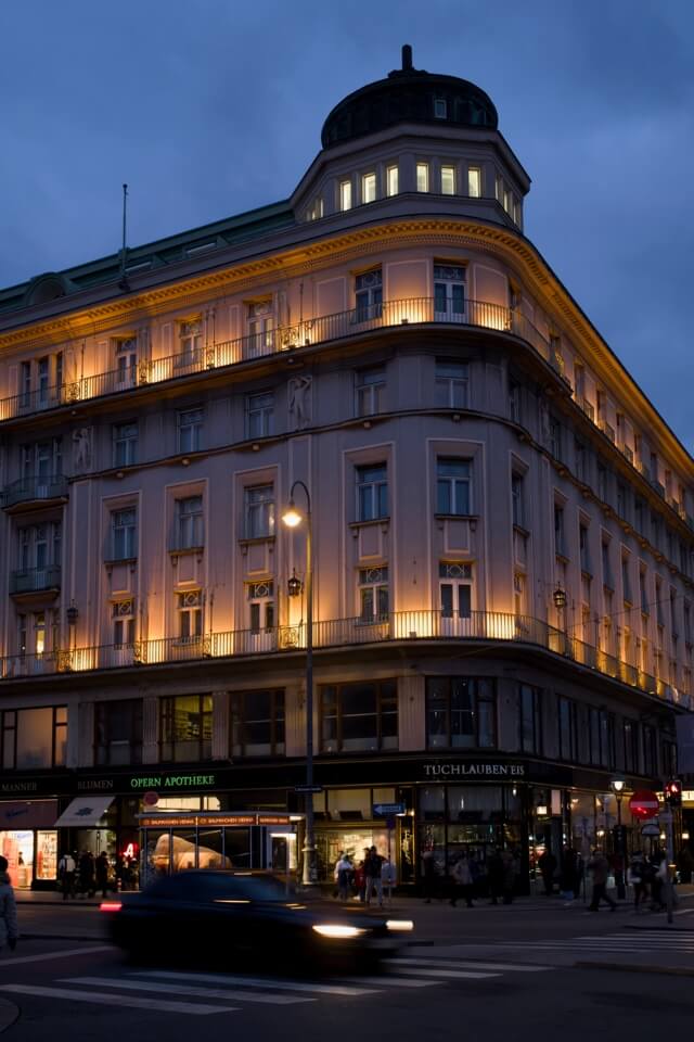 Ein beleuchtetes Eckhaus mit der Opern-Apotheke im Erdgeschoss in Wien bei Dämmerung.