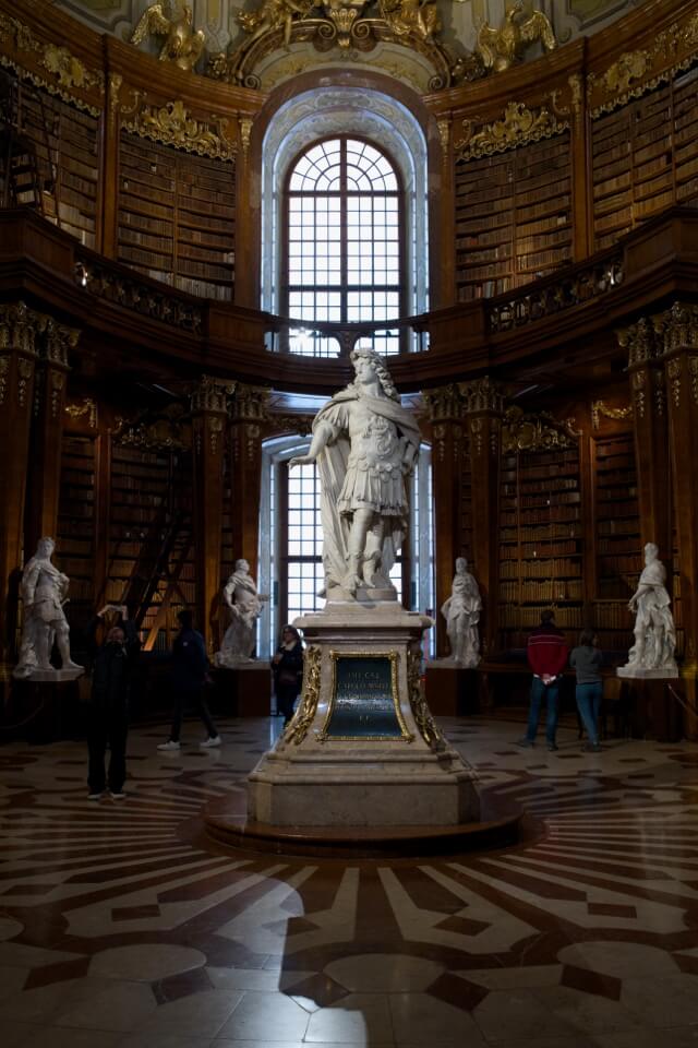 Der Prunksaal der österreichischen Nationalbibliothek mit einer großen Marmorstatue und historischen Bücherregalen.