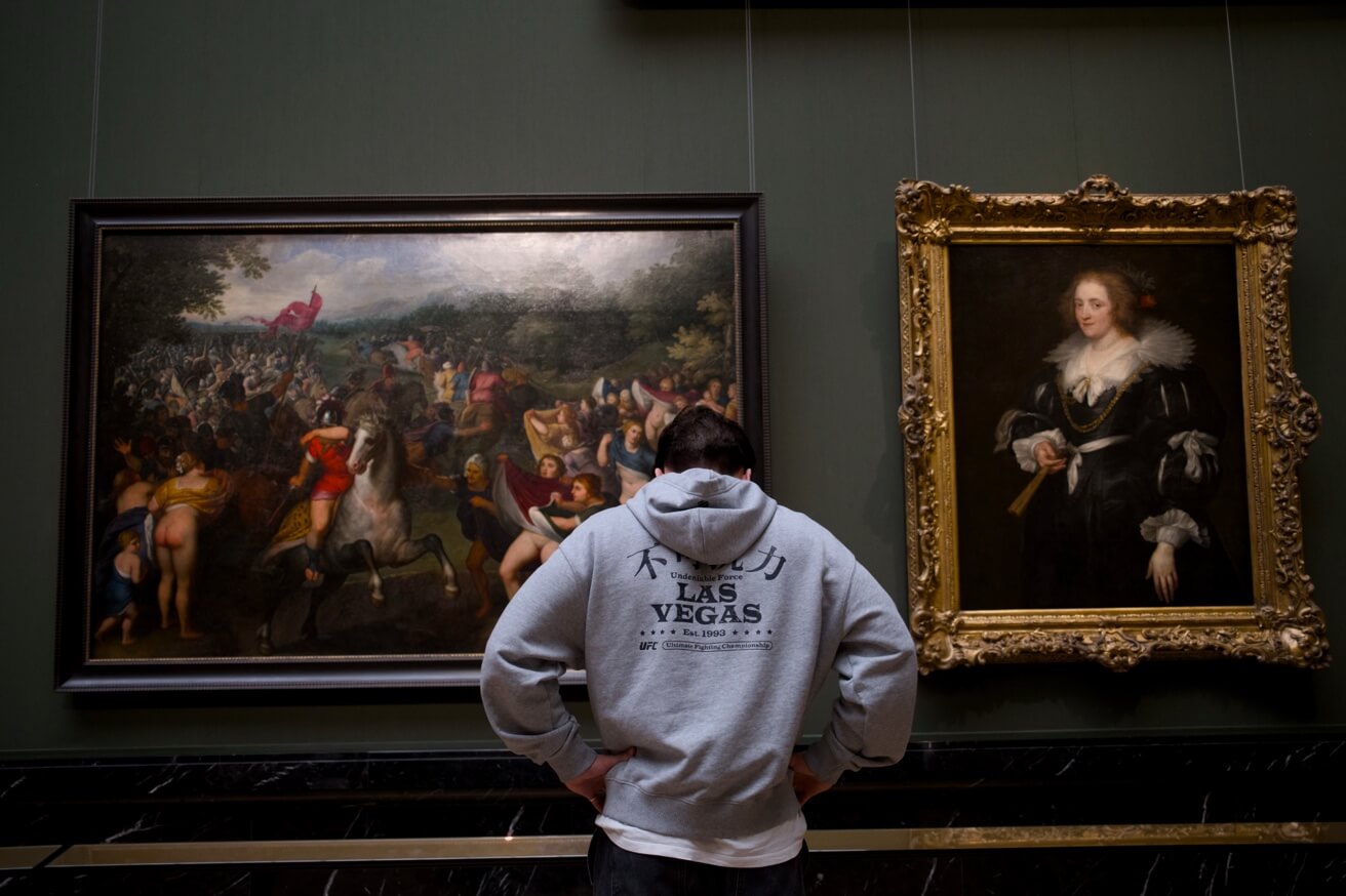 Ein Mann im grauen Kapuzenpullover betrachtet von hinten zwei große Ölgemälde in einem Museum.