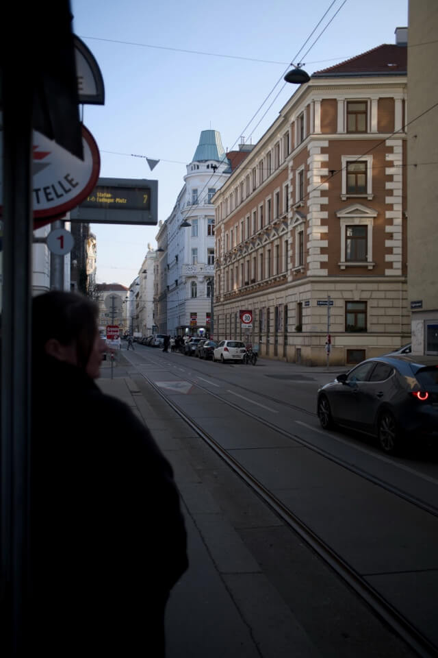Eine Person wartet an einer Haltestelle in einer typischen Wiener Straße mit Altbauten und Straßenbahnschienen.