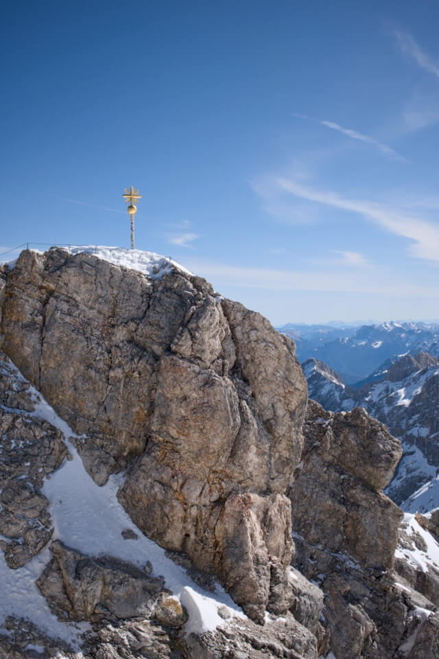 Das vergoldete Gipfelkreuz steht auf dem schneebedeckten Felsen der Zugspitze vor blauem Himmel.