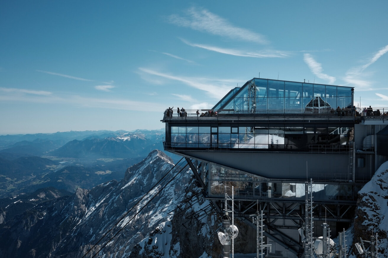 Besucher stehen auf der Aussichtsterrasse der modernen Gipfelstation der Zugspitze und blicken auf die Alpen.