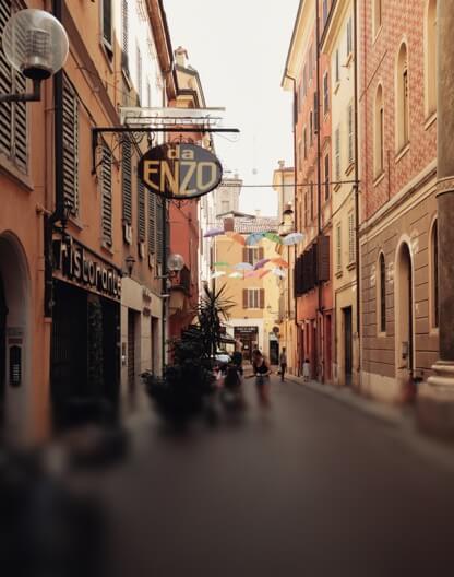 Eine enge Gasse in Italien mit dem Schild des Restaurants 'da Enzo' und bunten Regenschirmen.