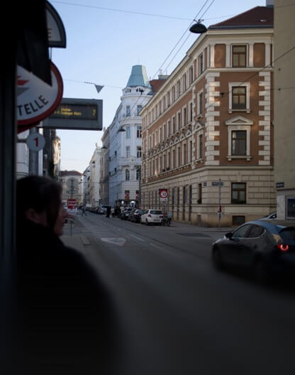 Eine Person wartet an einer Haltestelle in einer Wiener Straße mit Altbauten und Straßenbahnschienen.